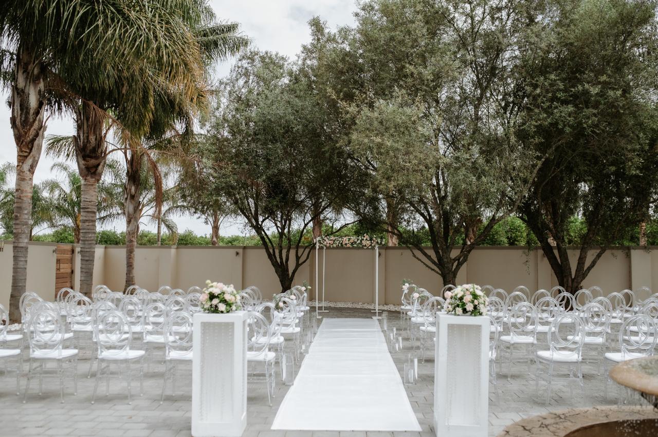 Outdoor white wedding ceremony with ghost chairs, white aisle runner and floral arch